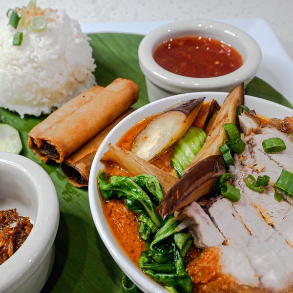 Filipino Kare-Kare with crispy pork belly, vegetables, rice, lumpia, and dipping sauces on a banana leaf.
