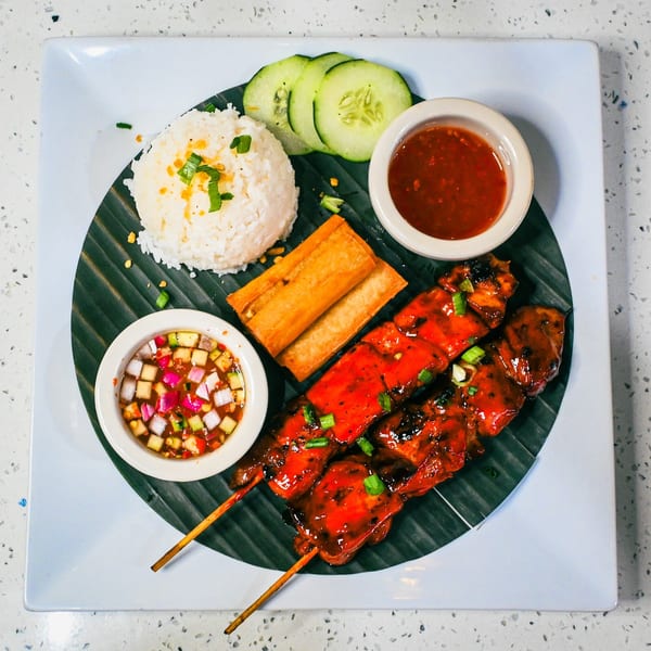 Grilled chicken skewers with rice, spring rolls, cucumber, and two dipping sauces on a banana leaf.